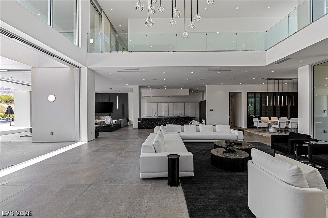This expansive, open-concept living area features a modern aesthetic with a large white sectional sofa centered on a dark area rug. The space is defined by high ceilings, a dramatic multi-pendant light fixture, and large glass walls that slide open to connect the interior with the outdoor patio. In the background, a sleek kitchen with a dark stone island and a formal dining area complete the sophisticated, airy layout.