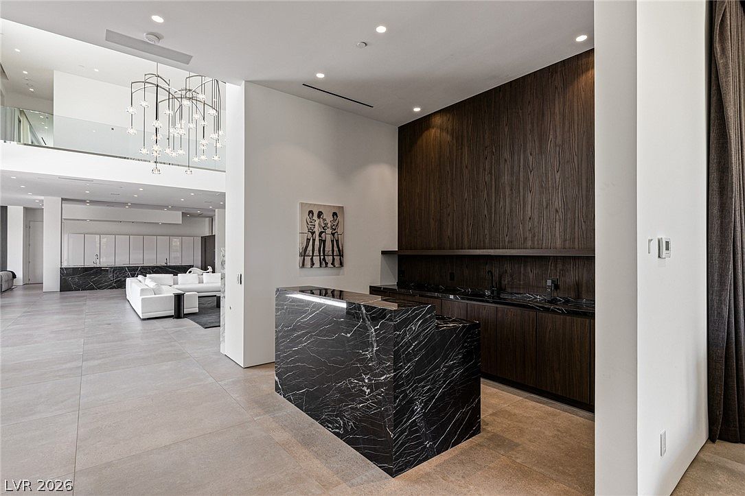 This modern, open-concept living area features a striking wet bar with a dark stone counter and matching cabinetry set against a dark wood-paneled wall. The space flows into a bright, expansive living room with large white sofas and a dramatic, multi-tiered chandelier hanging from the high ceiling. The neutral color palette and clean lines create a sophisticated, high-end atmosphere.