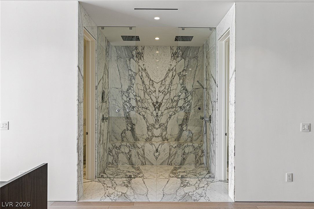 This luxurious walk-in shower features dramatic, book-matched patterned stone slabs that create a striking visual focal point. The space is enclosed by clear glass panels and includes dual overhead rain shower heads, a built-in stone bench, and a handheld shower attachment. The minimalist design is framed by white walls and light-toned wood flooring, emphasizing a high-end, contemporary aesthetic.