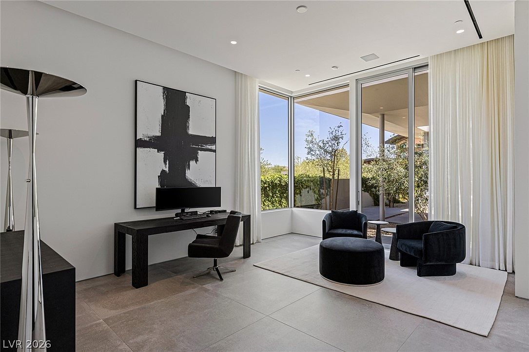 This modern home office features a minimalist aesthetic with a dark wood desk, a sleek black office chair, and a large abstract art piece on the wall. The room is anchored by a seating area with two black armchairs and a matching ottoman on a light-colored rug, all positioned near expansive floor-to-ceiling windows that provide abundant natural light and views of the outdoor greenery. The space is finished with neutral large-format floor tiles and recessed lighting, creating a sophisticated and professional atmosphere.