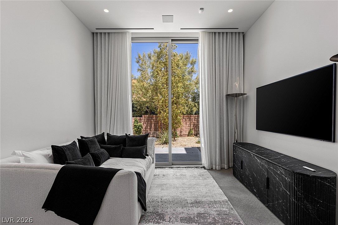 This contemporary living room features a plush, light-colored sectional sofa adorned with dark accent pillows, positioned across from a sleek, dark stone-textured media console. A large television is mounted on the wall above the console, while floor-to-ceiling glass sliding doors provide a clear view of the landscaped backyard. The space is finished with neutral carpeting and soft, light-colored curtains, creating a calm and sophisticated atmosphere.