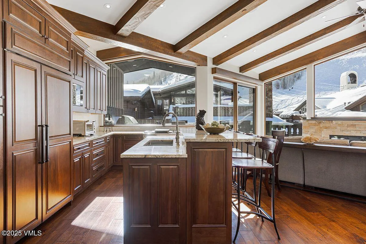 This is a warm and inviting kitchen featuring dark wood cabinetry, granite countertops, and stainless steel appliances. The kitchen island offers bar seating, and the large windows provide ample natural light and picturesque views of the snowy exterior. The exposed beam ceiling adds a touch of rustic charm to the space.