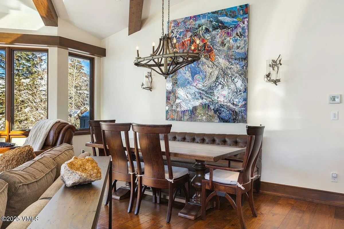 This image showcases an elegant dining room, featuring a long wooden table with seating for several people. The room is illuminated by a unique chandelier and supported by sconces on the wall, enhancing the sophisticated decor. An abstract artwork adorns the wall, adding a splash of color and artistic flair to the space. Hardwood floors and wood trim detail complete the room's design, suggesting warmth and luxury.