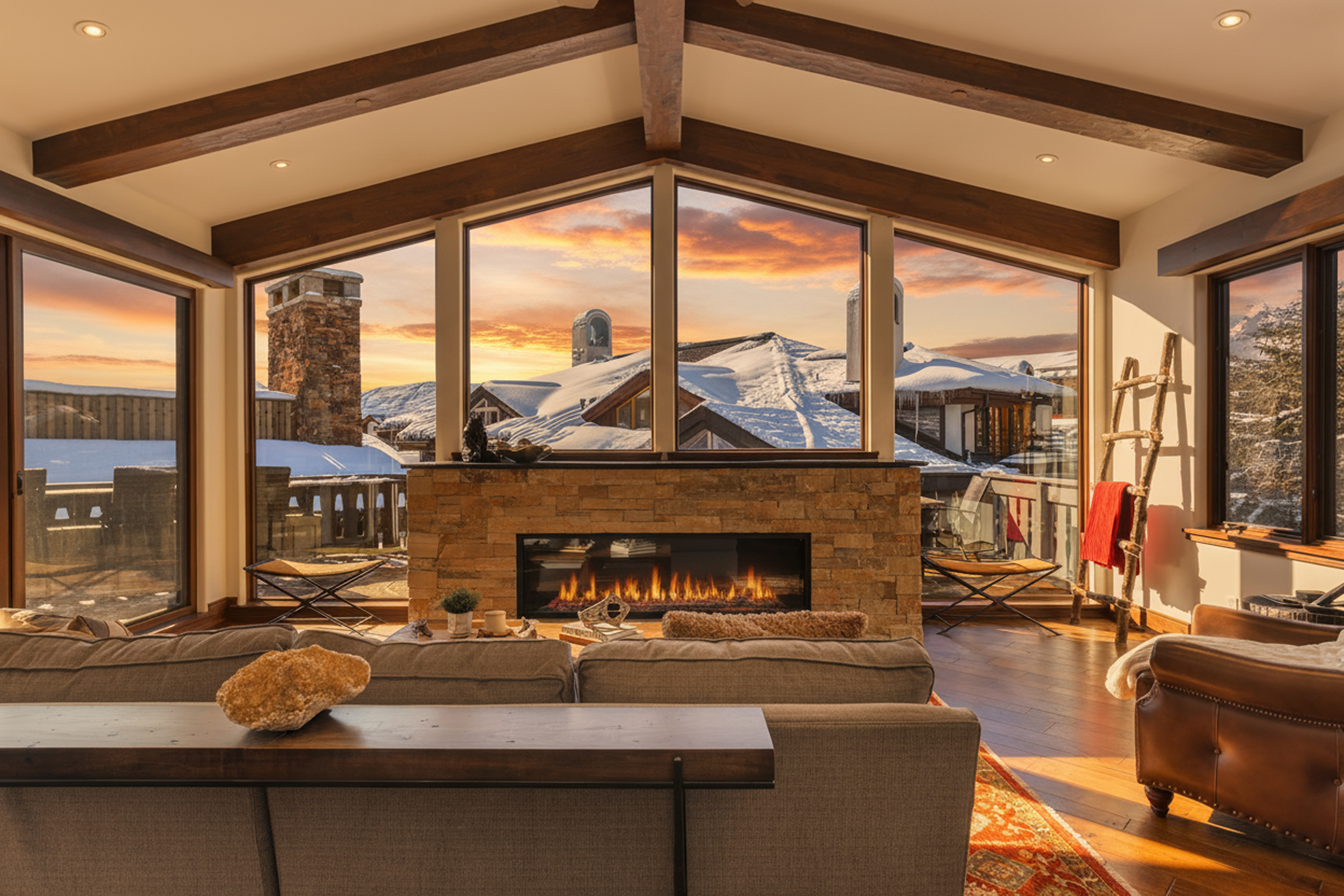 This interior shot showcases a luxurious living room featuring a modern fireplace with a stone surround and panoramic windows offering a breathtaking view of a snow-covered landscape at sunset. The room is elegantly furnished with comfortable seating and wood beams accentuating the ceiling, adding warmth and character. The design creates a cozy and inviting atmosphere, emphasizing the property's appeal as a high-end mountain retreat.