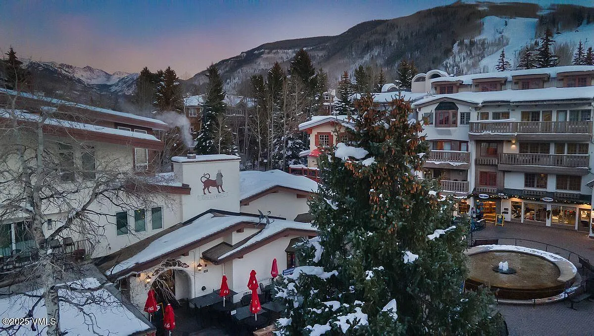 This aerial view shows a charming ski resort town nestled amidst snow-capped mountains and evergreen trees. The architectural style is alpine, with buildings featuring steeply pitched roofs dusted with snow. A central plaza features a fountain and outdoor dining, creating a lively and inviting atmosphere.