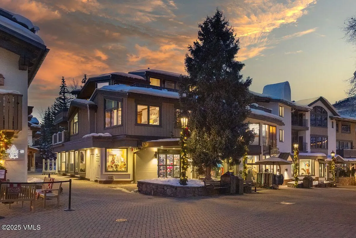 This image showcases the front exterior of a commercial building with architectural details suitable for a retail or office space. The building features a mix of wood and stucco finishes, with snow-covered roofs and windows that emit a warm interior glow. The surrounding pedestrian area is paved and decorated with street lamps and benches, creating a welcoming and picturesque atmosphere.