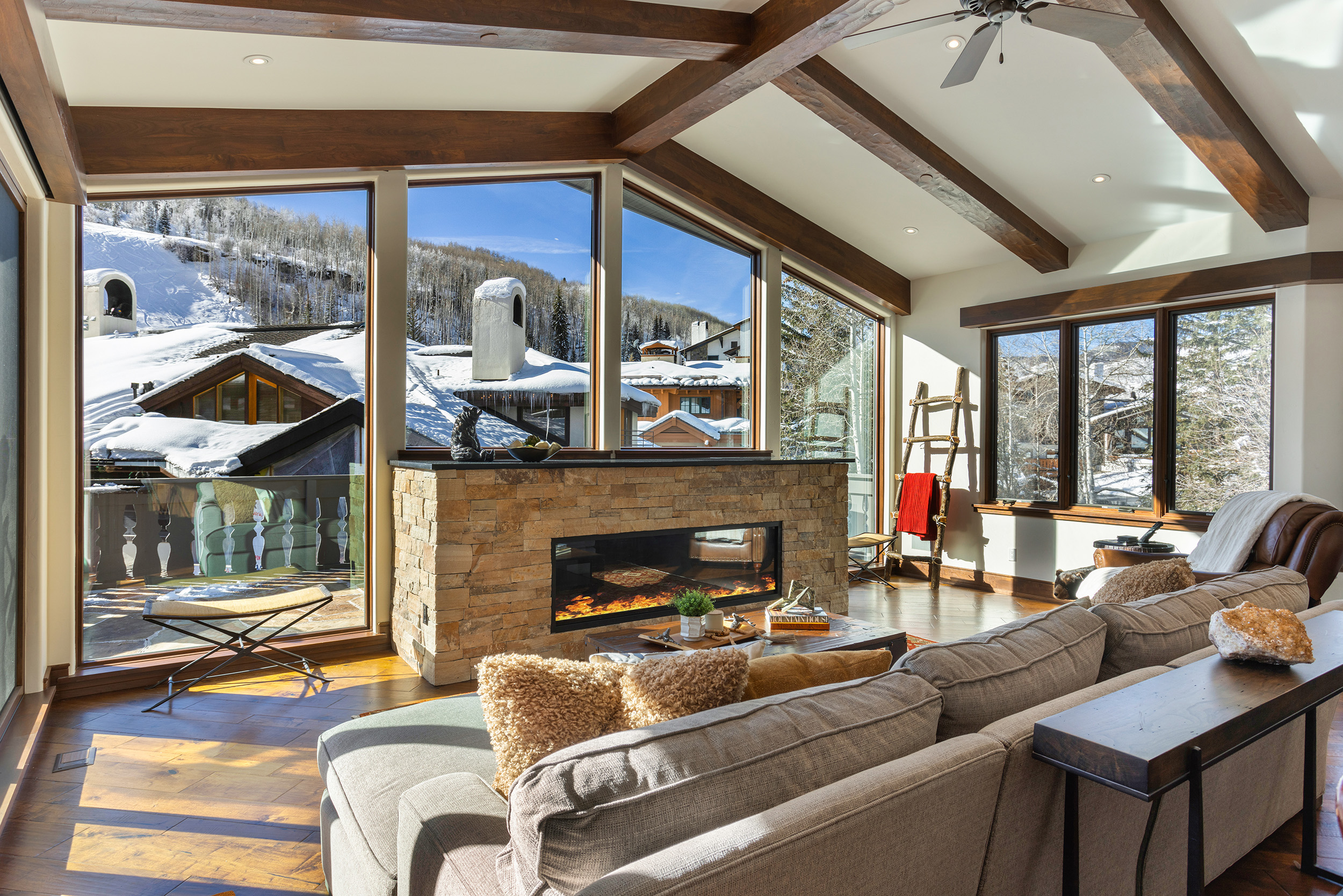 The image showcases a cozy living room with a modern fireplace and stunning views of a snow-covered landscape. The room features exposed wooden beams, hardwood floors, and a comfortable-looking sectional sofa, creating a warm and inviting atmosphere. Large windows maximize the natural light and offer panoramic vistas, highlighting the property's connection to its beautiful surroundings.