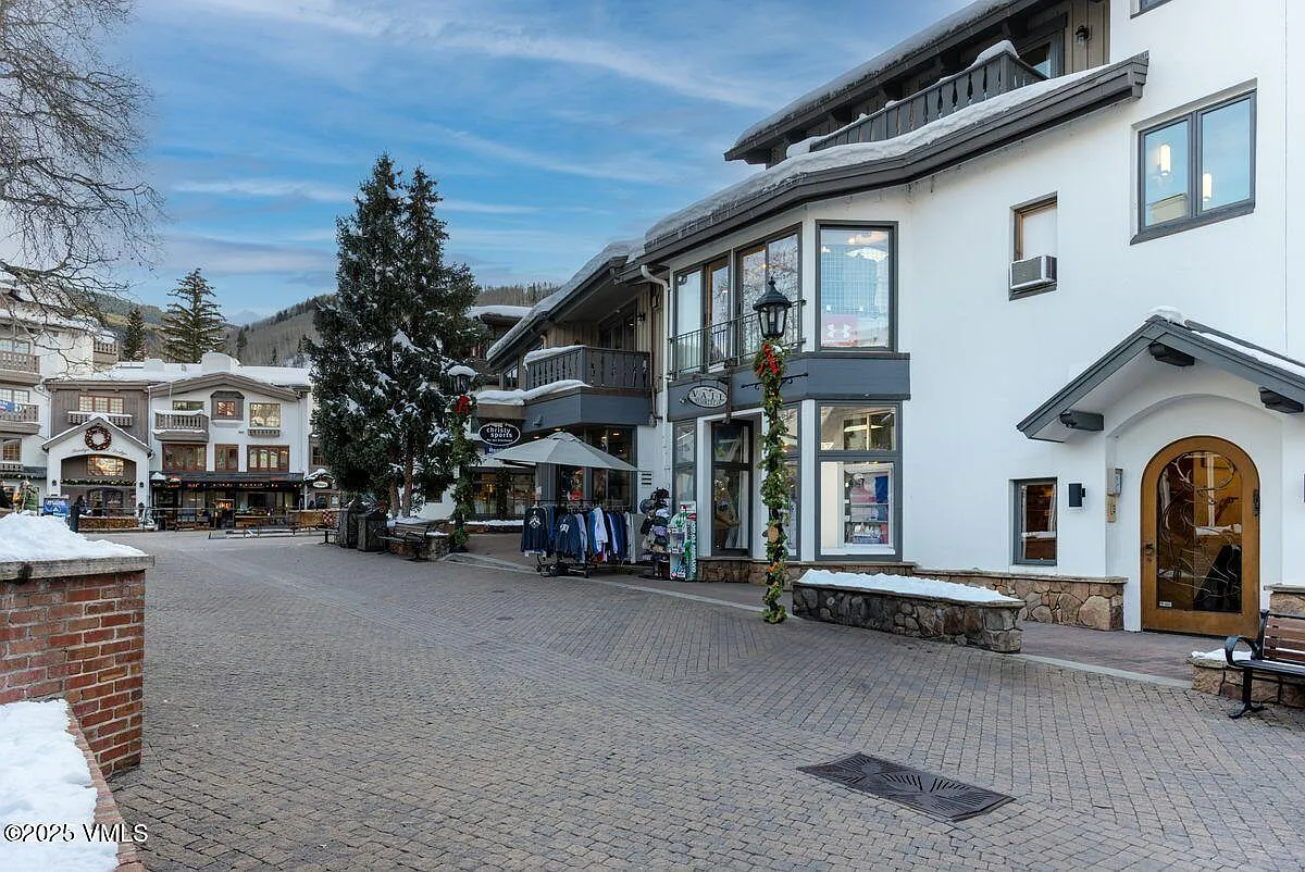This is a front view of a commercial building with retail storefronts. The architecture features a mix of stone and white siding, with distinctive window designs and decorative entryways. Snow accents the roofs and sidewalks, lending a charming, wintry aesthetic.