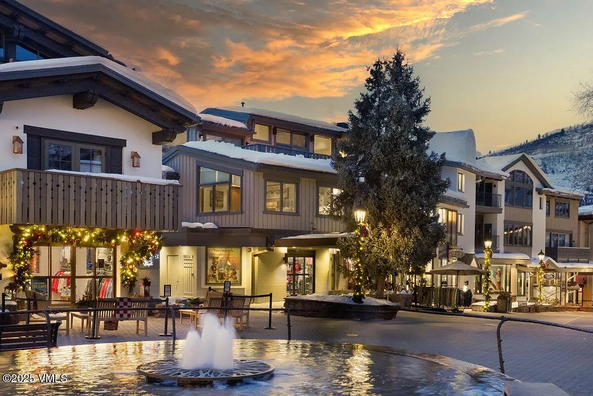 This image showcases the front view of multi-story buildings in what appears to be a village setting, possibly a ski resort town. The architecture includes features like balconies and decorative lighting, with a notable water fountain in the foreground. The photo is taken during twilight, with a cloudy sky adding a vibrant backdrop, creating an inviting and pleasant atmosphere.