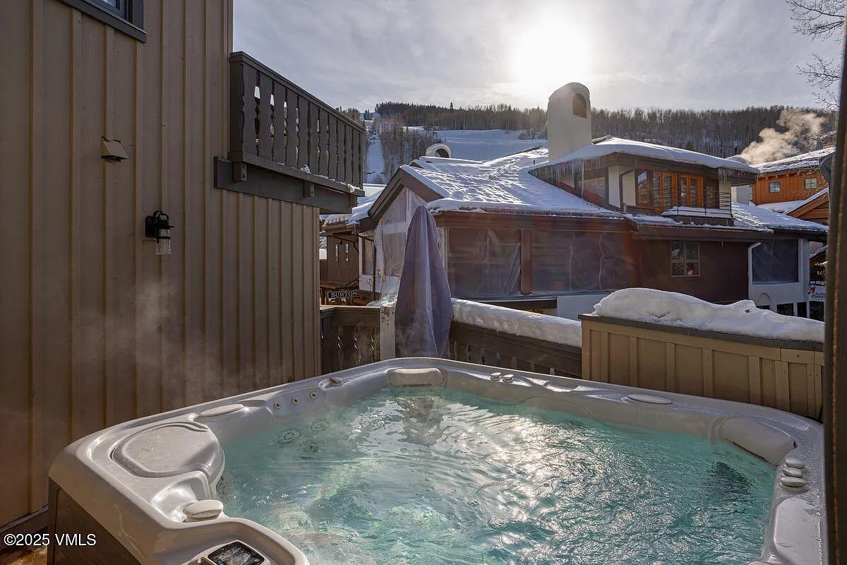 This image showcases a luxurious outdoor spa area, featuring a hot tub filled with clear, bubbling water. The surrounding landscape includes snow-covered roofs of nearby buildings, suggesting a mountain or cold climate. The setting implies a relaxing and private amenity within a residential property.