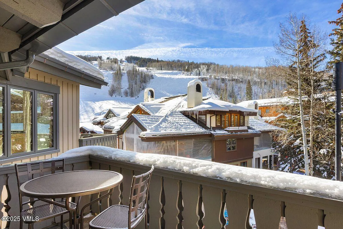 This image showcases a charming balcony with a view of a snowy landscape. The balcony features a table and chairs, inviting relaxation. The backdrop includes snow-covered rooftops of nearby buildings and a scenic hillside, creating a cozy and picturesque atmosphere, highlighting an outdoor living space.