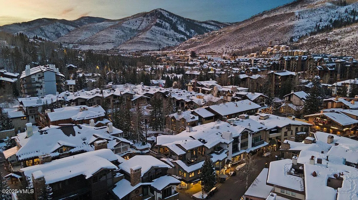 This aerial view showcases a luxurious mountain resort community blanketed in snow. Multiple buildings, likely condos or townhomes, feature snow-covered roofs, with the mountainous backdrop adding to the scenic appeal. The lighting suggests either dawn or dusk, contributing to the serene and high-end ambiance of this real estate offering.