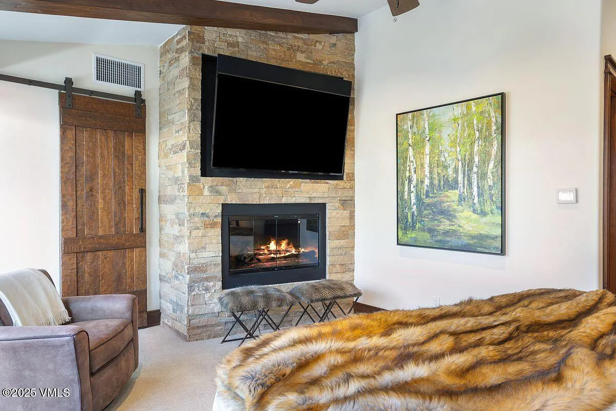 This is an inviting primary bedroom featuring a stone fireplace with a mounted TV, a unique rustic sliding barn door, and a cozy seating area. The room exudes a lodge-like atmosphere with its wooden touches and a luxurious fur throw on the bed, suggesting a comfortable and relaxing space. The artwork on the wall adds a touch of nature and complements the overall ambiance.