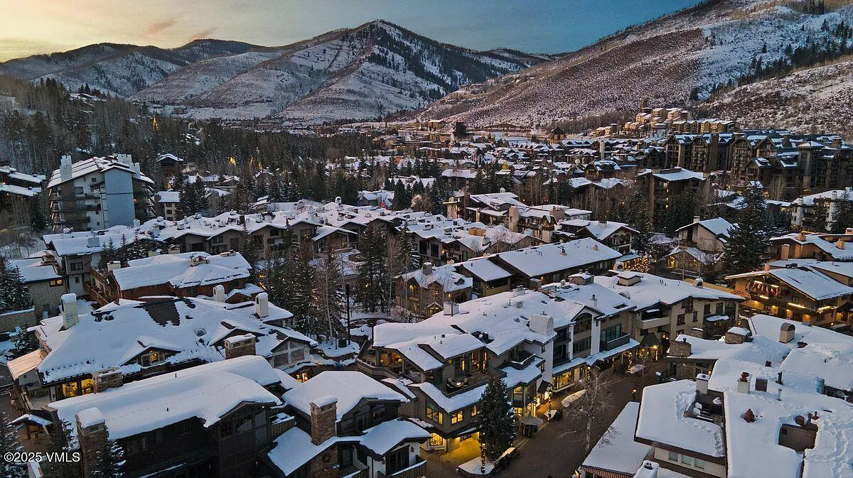 This is an aerial view of a snow-covered residential area nestled in a mountainous landscape. The buildings feature varying architectural styles with snow-covered roofs and exteriors. The setting conveys a sense of a cozy, winter retreat, highlighting the property's location within a picturesque environment.