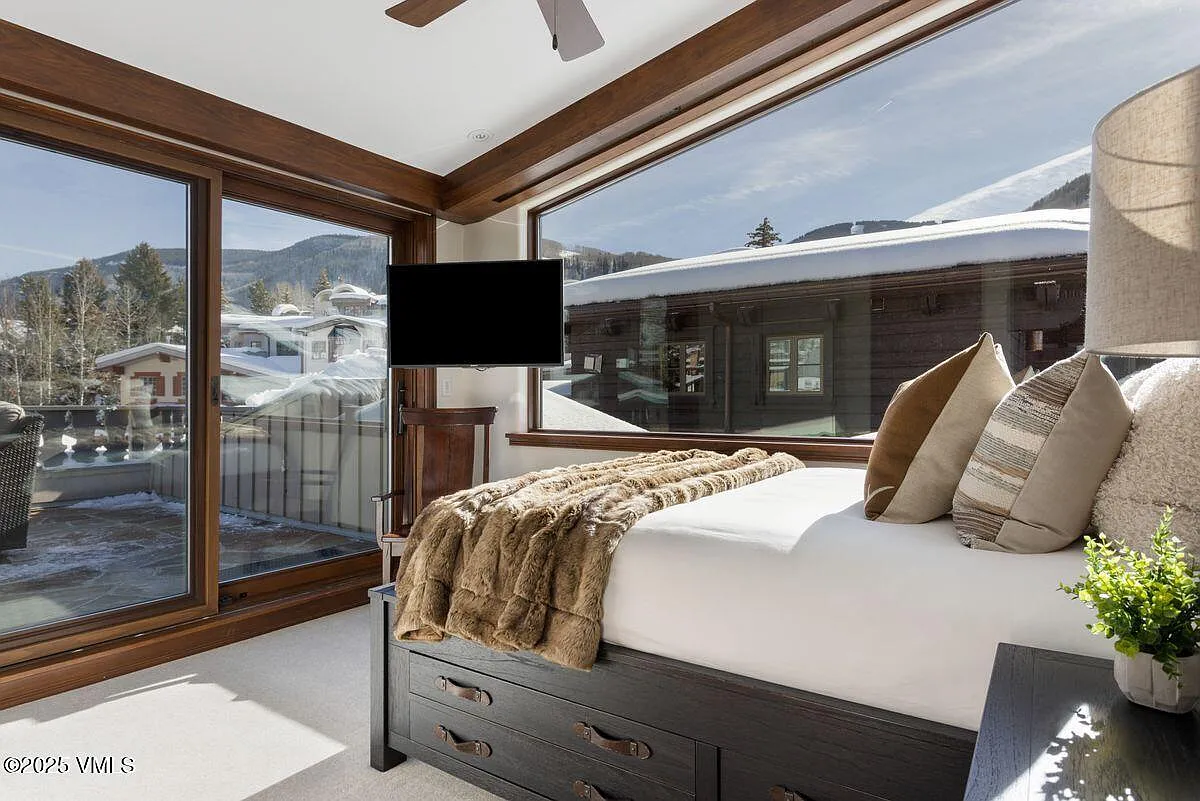 The image showcases a stylish primary bedroom featuring a bed with built-in storage and plush, comfortable bedding. A large window provides a scenic view of a snow-covered landscape. The room incorporates wood trim, which adds warmth and character, creating a cozy and inviting atmosphere.