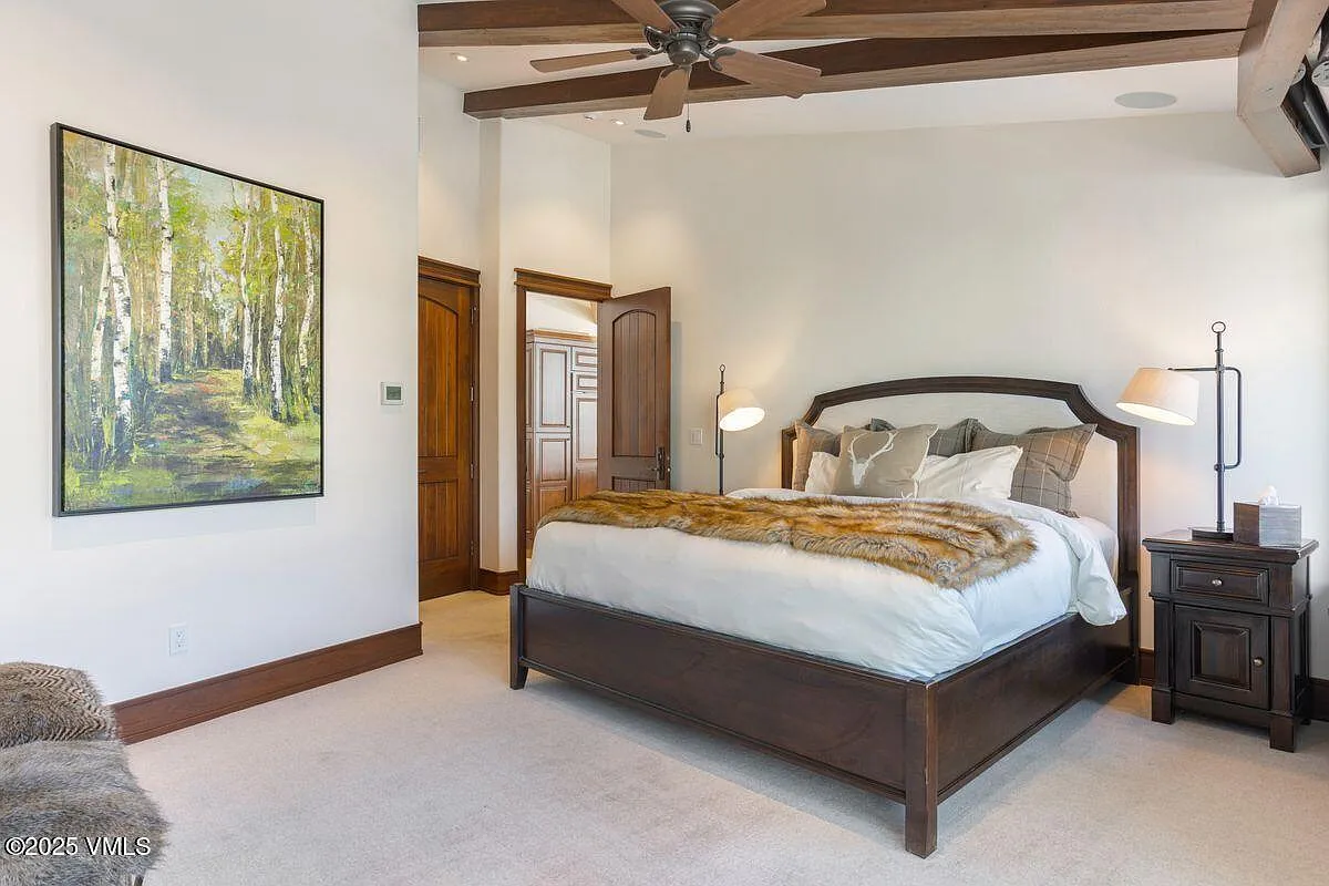 This is a primary bedroom featuring a queen bed with a fur throw blanket, a dark wooden nightstand with a lamp, and a large painting on the wall. The ceiling has exposed wooden beams and a ceiling fan. The room is decorated in a neutral color palette with wooden accents, lending it a cozy and inviting atmosphere.