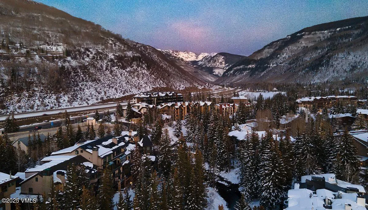This is an aerial view showcasing a mountain resort town in winter. The scene features snow-covered buildings nestled among evergreen trees with snow-capped mountains in the background. The image highlights the scenic beauty and resort-style living available in this location.
