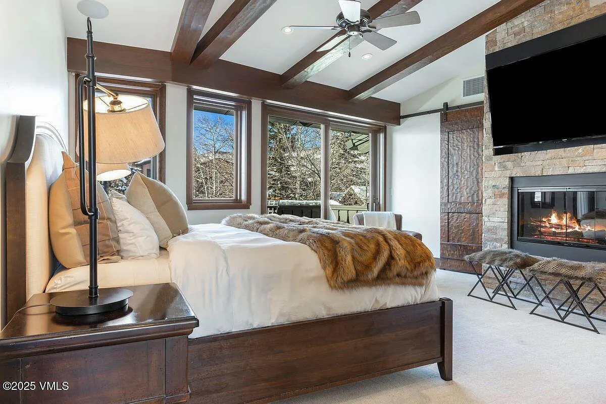 This is a primary bedroom featuring a stylish and comfortable design. The bed is adorned with plush bedding and a faux fur throw, creating a cozy atmosphere. The room also includes a stone fireplace with a mounted television above, adding a luxurious touch.
