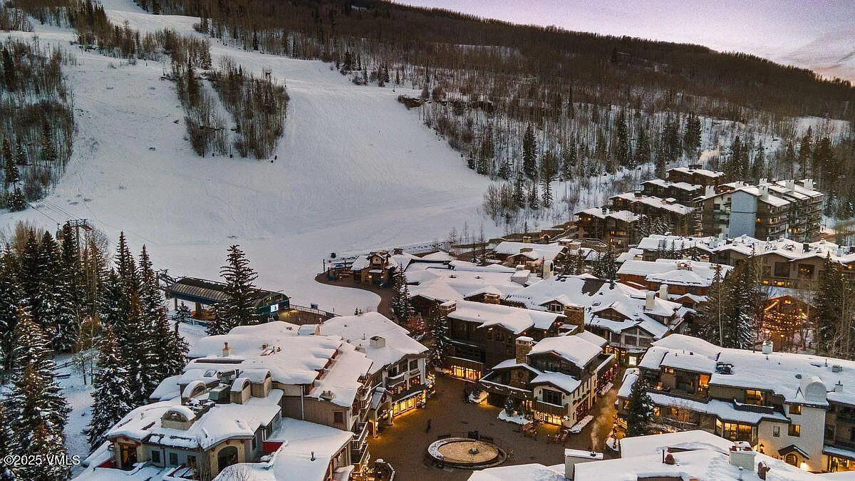 This is an aerial view of a charming, snow-covered village nestled in a mountainous setting. The architecture consists of multi-story buildings with gabled roofs, heavily blanketed in snow, creating a picturesque winter scene. The surrounding landscape features a ski slope and dense evergreen forests, likely appealing to potential buyers interested in a ski resort property.