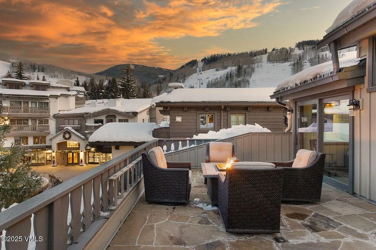 This image showcases a cozy outdoor patio space with wicker seating arranged around a fire pit, perfect for enjoying evenings with a view. The stone-tiled patio extends from a building with sliding glass doors, suggesting easy indoor-outdoor living. Snow-capped rooftops and mountain scenery create a serene, upscale alpine setting.