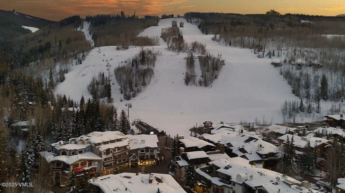 This aerial view captures a luxurious ski resort town nestled at the base of a snow-covered mountain. The buildings are densely packed with snow-covered roofs, and ski slopes provide a backdrop. The setting evokes a sense of winter recreation and high-end real estate.