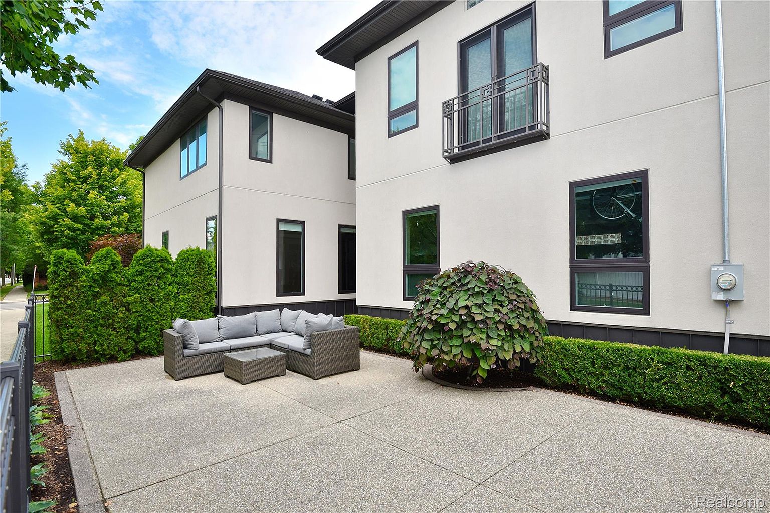 This image showcases a modern patio area situated between two sections of a contemporary home. The patio features a gray sectional sofa and coffee table, perfect for outdoor lounging. Lush greenery surrounds the space, providing privacy and enhancing the aesthetic appeal, while the clean lines of the building and the well-maintained patio surface contribute to a sophisticated outdoor living space.
