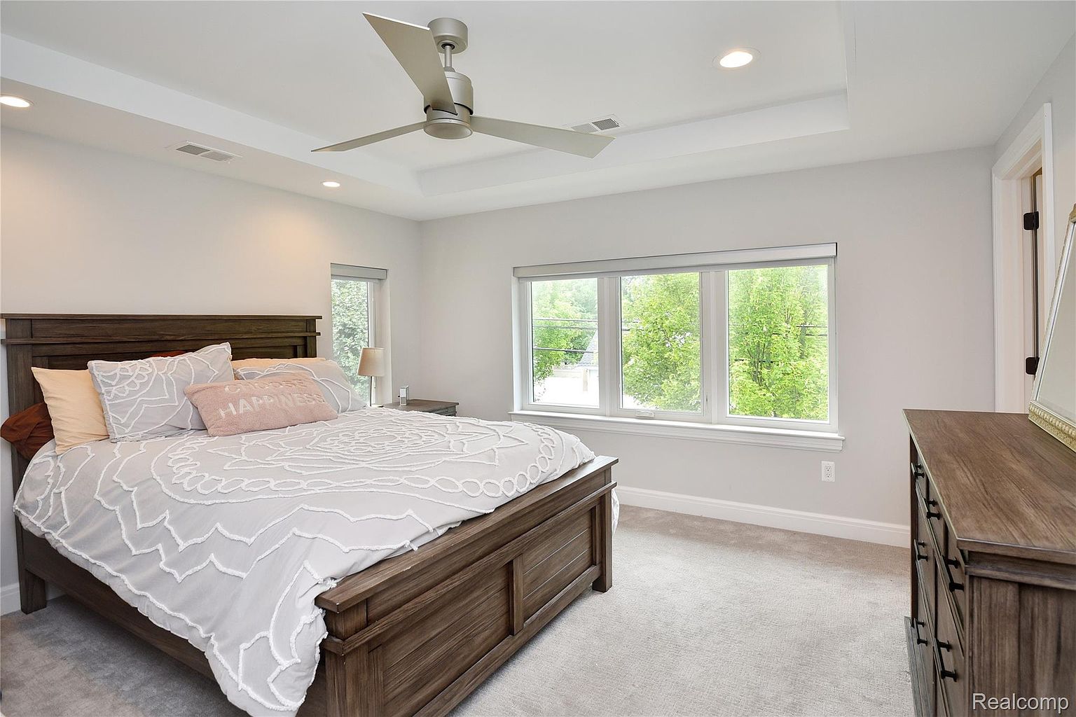 This is a well-lit primary bedroom featuring a large wooden bed with a white patterned duvet and several pillows, including one with the word 'HAPPINESS'. The room has a neutral color palette with white walls and light gray carpeting. A ceiling fan and recessed lighting provide illumination, and a bank of windows offers natural light and a view of greenery outside.