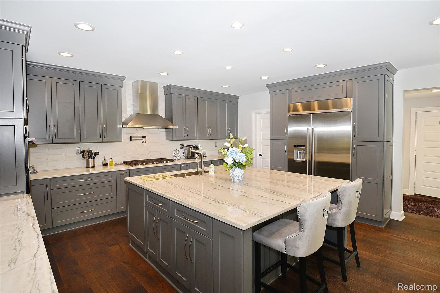 This is a well-lit kitchen featuring gray cabinetry, stainless steel appliances, and a large island with a marble countertop. The kitchen includes a stainless steel range hood and a white backsplash. The dark hardwood floors add warmth to the space, and the overall impression is modern and stylish.