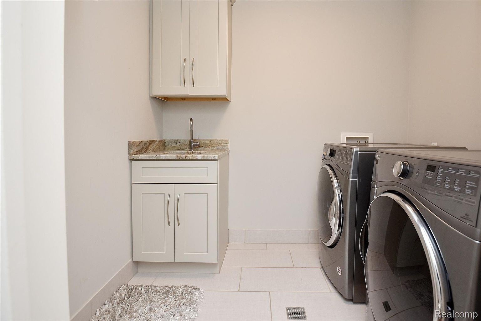 This is a well-organized laundry room featuring a sink with granite countertops and white cabinetry above and below. Modern, front-loading washer and dryer units are present, and the room is finished with light-colored tile flooring. The overall impression is clean and functional.