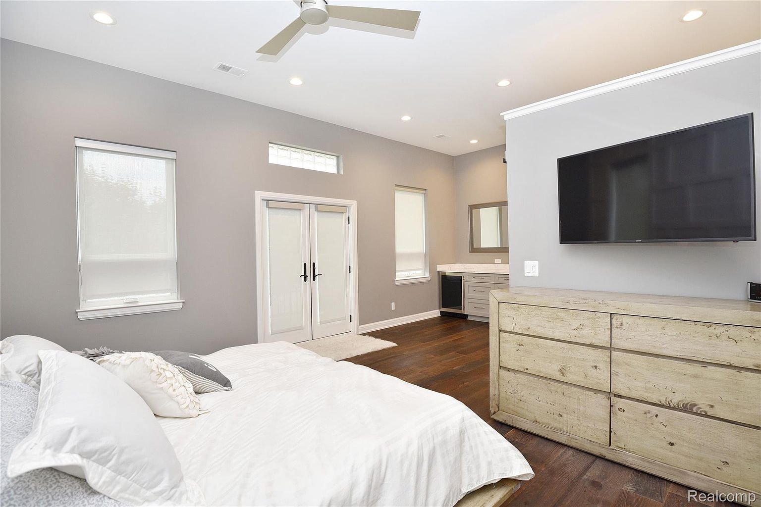 This is a primary bedroom featuring a large bed with white linens and decorative pillows. The room has a modern aesthetic with gray walls, dark hardwood floors, and white trim. A flat-screen TV is mounted on the wall above a light wood dresser, and there's a small bar area with a mirror and cabinetry in the background. The room is well-lit with natural light coming through the windows.