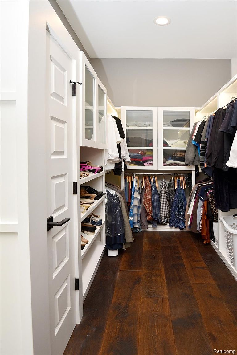 This is a well-organized walk-in closet featuring dark wood flooring and white shelving units. The closet is filled with clothing and shoes, and some of the shelving units have glass doors. The overall impression is one of neatness and efficient use of space.