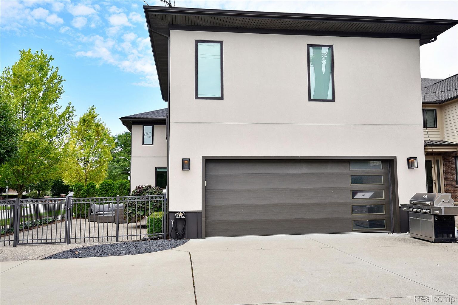 This image showcases the exterior of a modern home, focusing on the garage. The garage features a contemporary door with horizontal panels and glass inserts, complemented by sleek exterior lighting fixtures. A well-maintained concrete driveway leads to the garage, and a stainless steel grill is visible on the side, suggesting outdoor entertaining space.