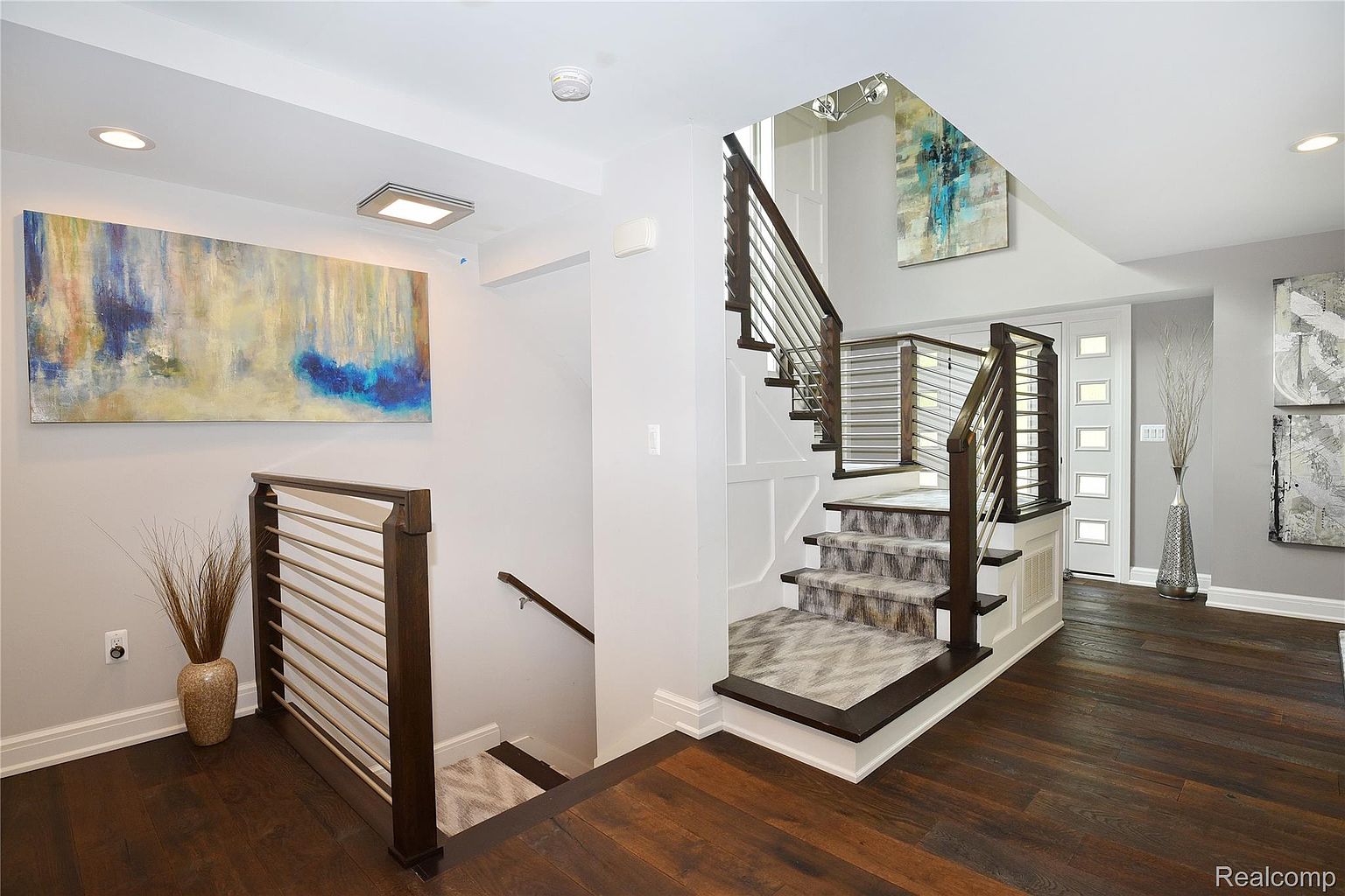 This interior shot showcases a modern hallway with a staircase. The staircase features dark wood steps with patterned carpet runners and stainless steel railings. Artwork adorns the walls, and the hardwood flooring adds warmth to the space. The overall impression is clean, bright, and stylish.