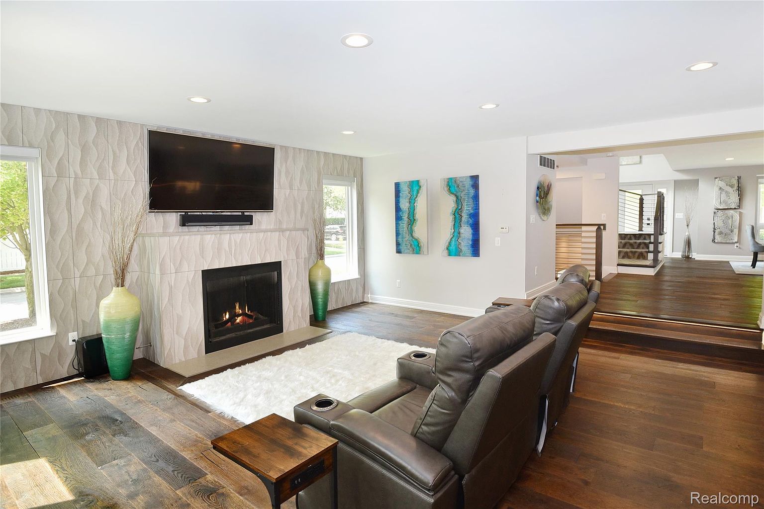 This is an interior shot of a living room featuring a modern fireplace with a large television mounted above it. The room has hardwood floors, a white shag rug, and a comfortable leather recliner. The space is well-lit with recessed lighting and natural light from the windows, creating a warm and inviting atmosphere.