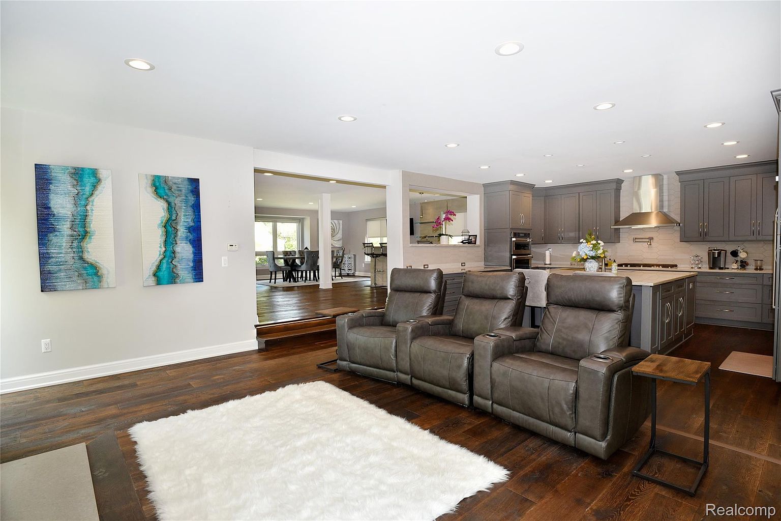 This interior shot showcases an open-concept living room that seamlessly transitions into a modern kitchen and dining area. The living room features a comfortable seating arrangement with leather recliners, a plush white rug, and stylish artwork. The kitchen boasts gray cabinetry, stainless steel appliances, and a spacious island, creating a cohesive and inviting living space.