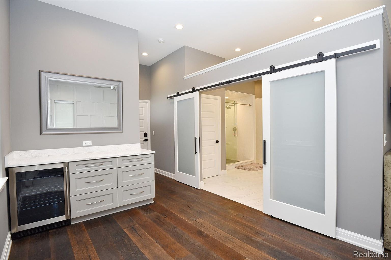 This is a well-lit primary bathroom featuring a modern design with gray walls and dark wood flooring. A gray cabinet with a white countertop and a built-in beverage cooler is on the left, and a large mirror hangs above it. Sliding barn doors with frosted glass panels lead to the shower area, adding a touch of elegance and privacy.