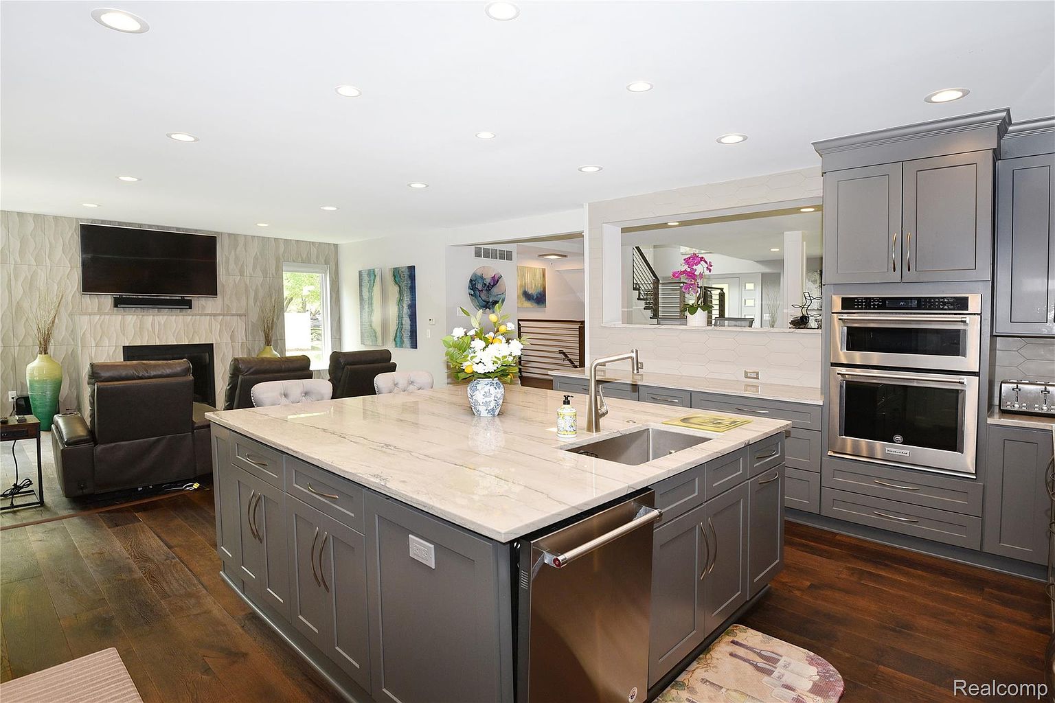 This is a well-lit, modern kitchen featuring gray cabinetry, stainless steel appliances, and a large island with a light-colored countertop. The kitchen opens to a living area with a fireplace and TV, creating an open-concept living space. The flooring is dark hardwood, adding warmth and contrast to the gray tones.
