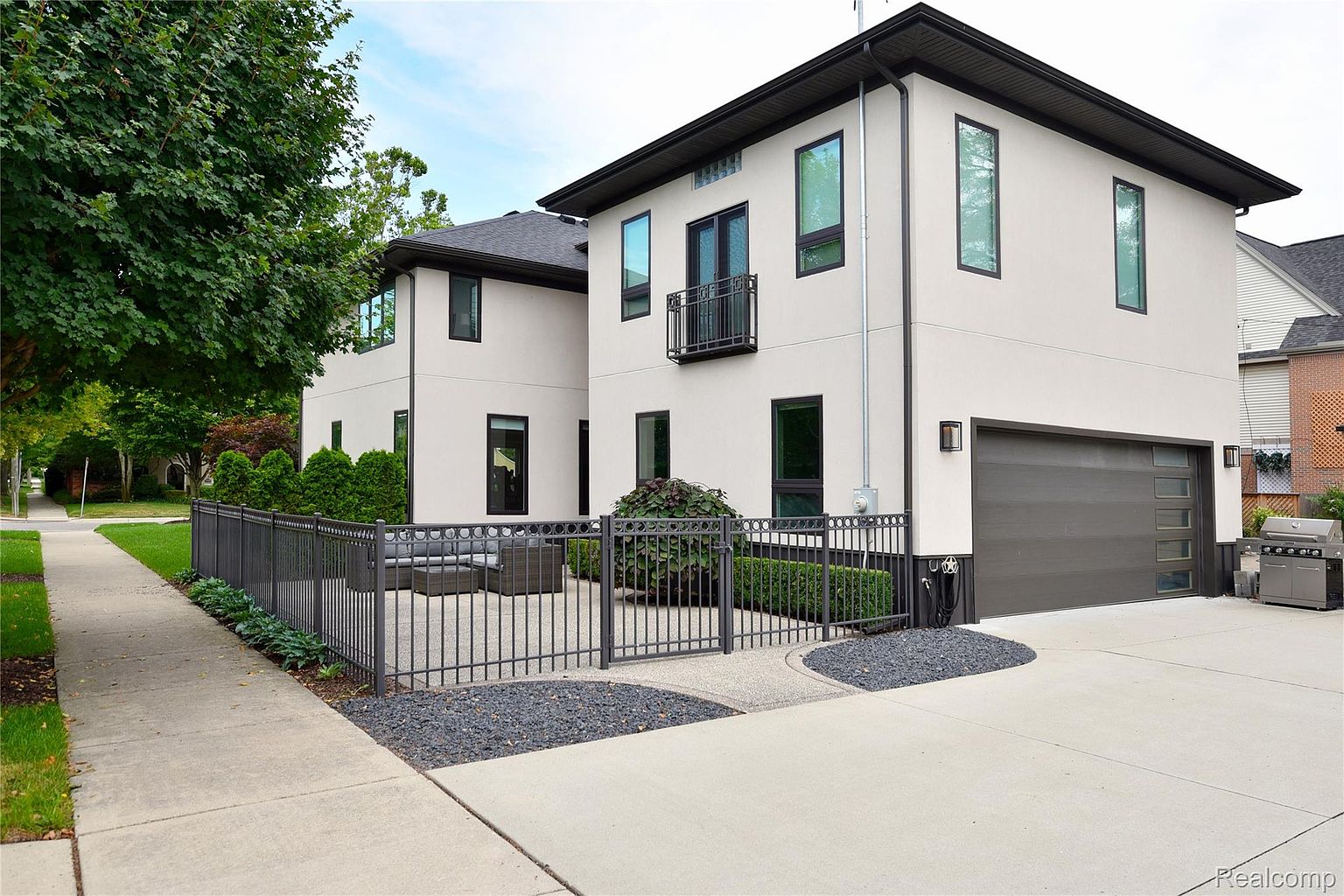 This is a side view of a modern two-story home with a light-colored stucco exterior and dark trim. The house features a dark garage door with windows, a small balcony, and a fenced-in patio area with outdoor seating. The landscaping includes manicured hedges and decorative gravel, adding to the property's curb appeal.
