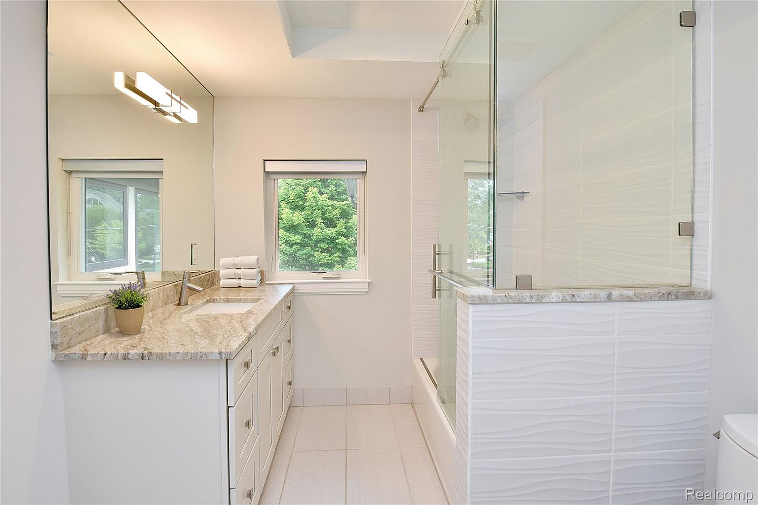 This is a bright and clean bathroom featuring a vanity with a light-colored granite countertop and white cabinets. A large mirror reflects the natural light coming in from the window above the sink. The shower area is enclosed with glass and features textured white tiles, adding a modern touch to the space.
