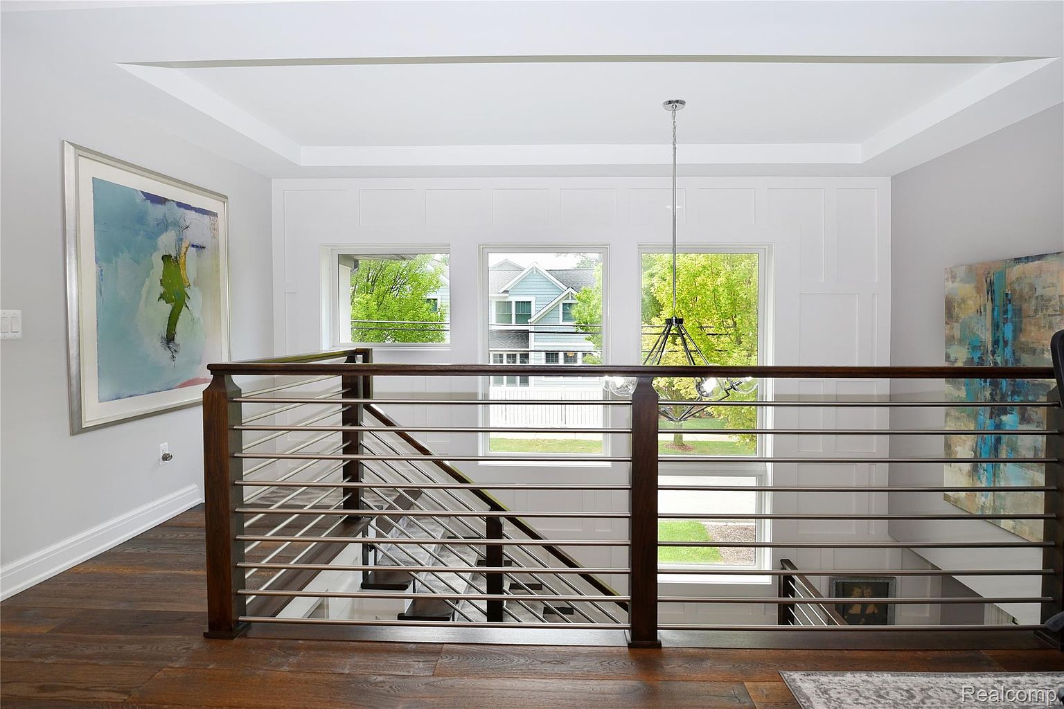This interior shot showcases a modern hallway and staircase with a focus on architectural details. The staircase features a dark wood and metal railing system, complemented by hardwood flooring. Natural light floods the space through a series of windows, highlighting the clean white walls and trim, and a chandelier hangs from the ceiling.