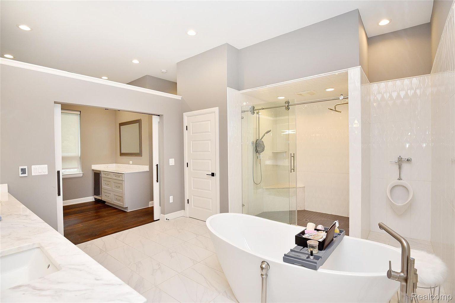 This is a luxurious primary bathroom featuring a freestanding white bathtub as a focal point, complemented by a glass-enclosed shower and a separate vanity area. The bathroom boasts a modern design with light gray walls, white tile flooring, and recessed lighting, creating a bright and spacious atmosphere. A unique feature is the inclusion of a urinal, adding a touch of sophistication and convenience.