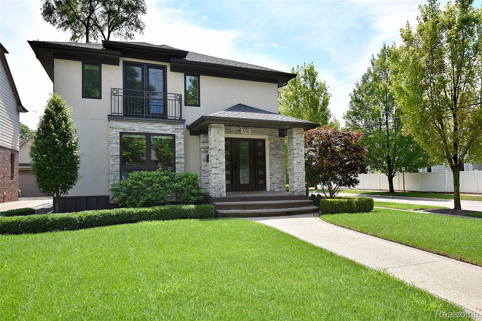 This is a front view of a modern two-story house with a light-colored stucco exterior and stone accents around the entryway and lower windows. The house features a small balcony, a well-manicured lawn, and a paved walkway leading to the front door. The landscaping is neat and tidy, enhancing the curb appeal of the property.