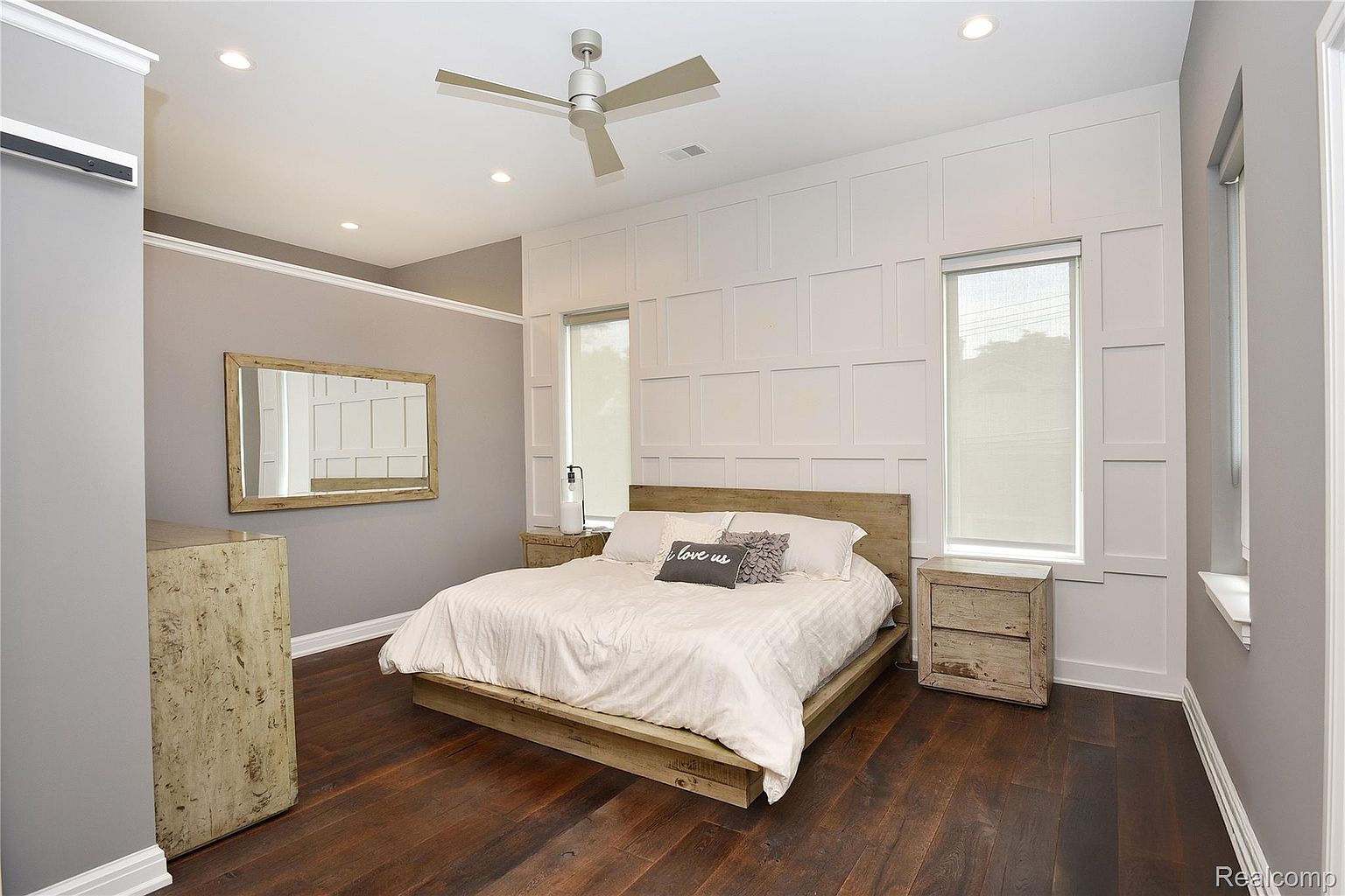 This is a primary bedroom featuring a wooden platform bed with a light-colored duvet and decorative pillows. The room has a neutral color palette with gray walls and white wainscoting, complemented by matching wooden nightstands and a dresser. Natural light streams in through the windows, creating a serene and inviting atmosphere.