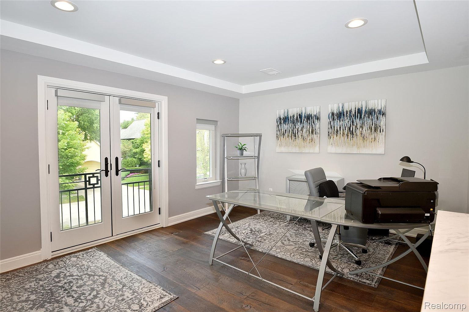 This is an interior shot of a home office featuring a glass-top desk, a gray office chair, and a printer. The room has a modern aesthetic with light gray walls, recessed lighting, and two abstract art pieces on the wall. Double doors lead to a balcony, providing natural light and a view of the outdoors.