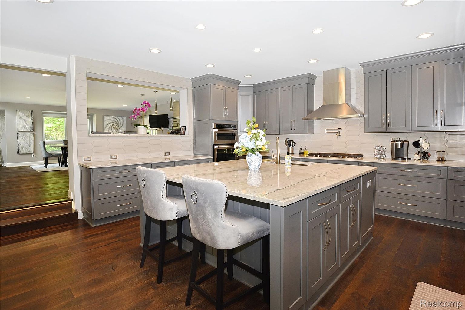 This is a well-lit kitchen featuring gray cabinetry, stainless steel appliances, and a large island with a light-colored countertop. The kitchen has a modern design with a pass-through to another room, and two bar stools are positioned at the island. The dark wood flooring adds warmth to the space.