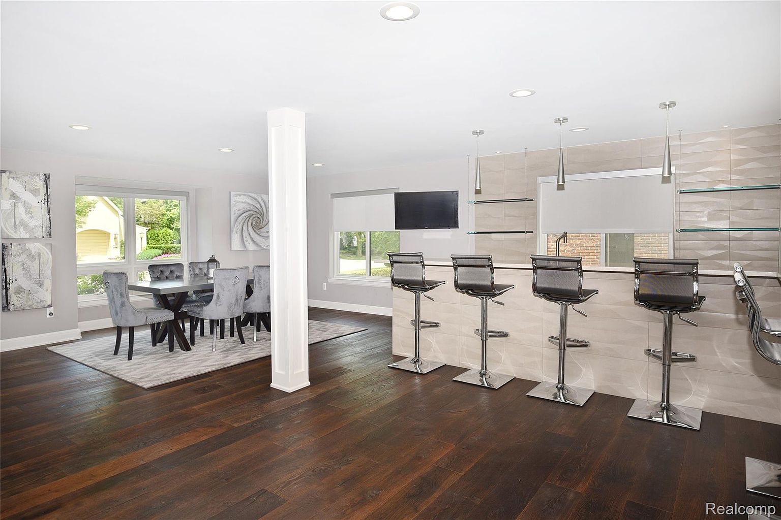 This interior shot showcases a dining area adjacent to a kitchen bar. The dining space features a modern table with six velvet chairs, complemented by artwork and a large window providing natural light. The kitchen bar includes seating and stylish pendant lighting, creating a seamless transition between the dining and kitchen areas.
