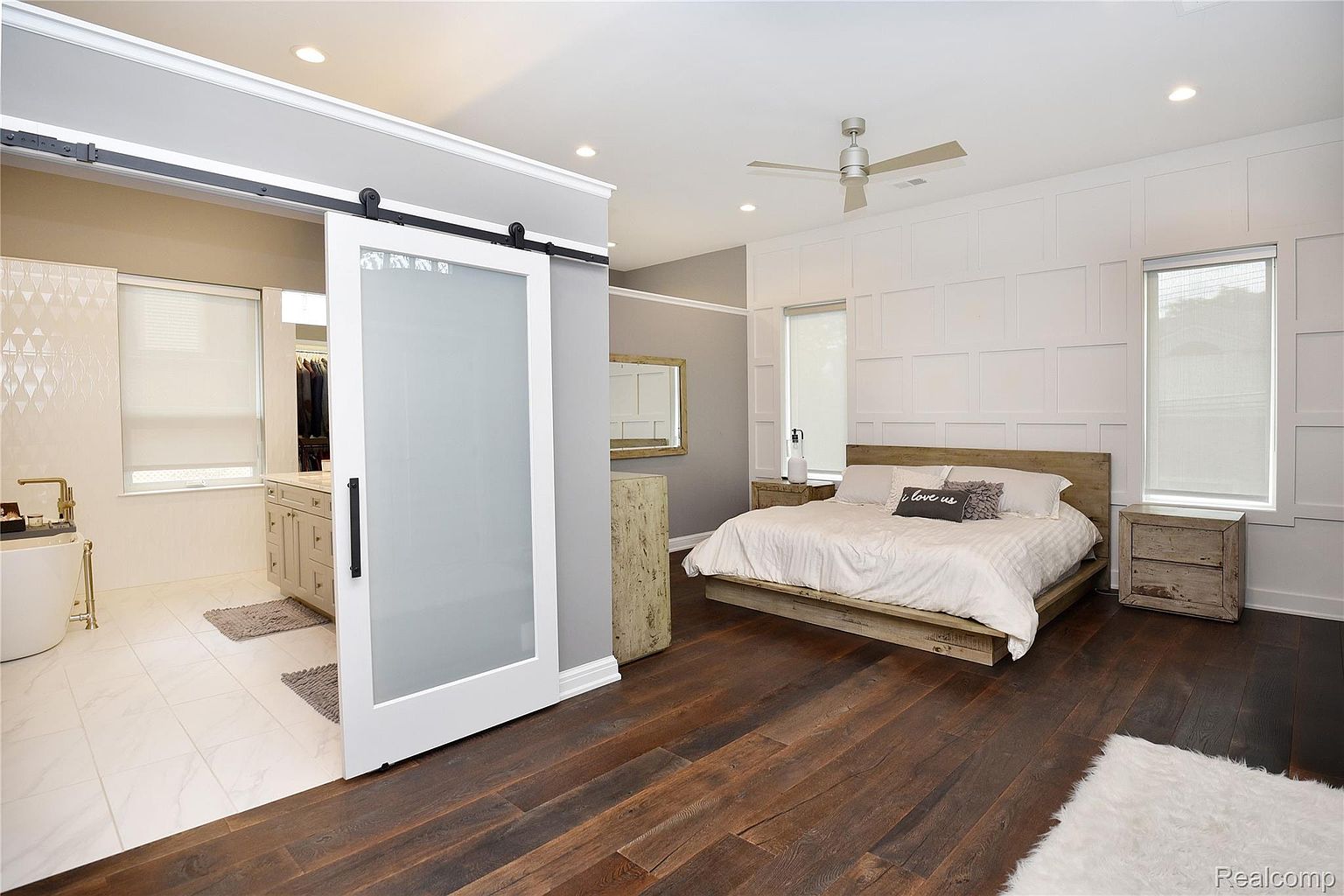 This is a primary bedroom featuring a modern farmhouse design with a rustic wood platform bed, white paneled accent wall, and dark hardwood floors. A sliding barn door leads to the primary bathroom, which has white tile flooring and a freestanding tub. The room is well-lit with natural light and recessed lighting, creating a serene and inviting atmosphere.