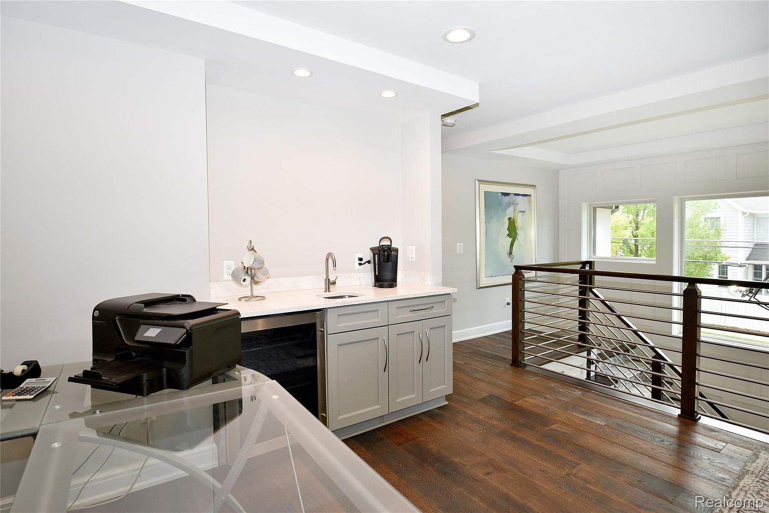 This interior shot showcases a modern office space featuring a wet bar with gray cabinetry and a white countertop, complete with a sink and beverage cooler. A printer sits on a glass desk, and the room transitions to a hallway with a wooden floor and a railing with horizontal metal bars. The overall impression is clean, functional, and stylish.