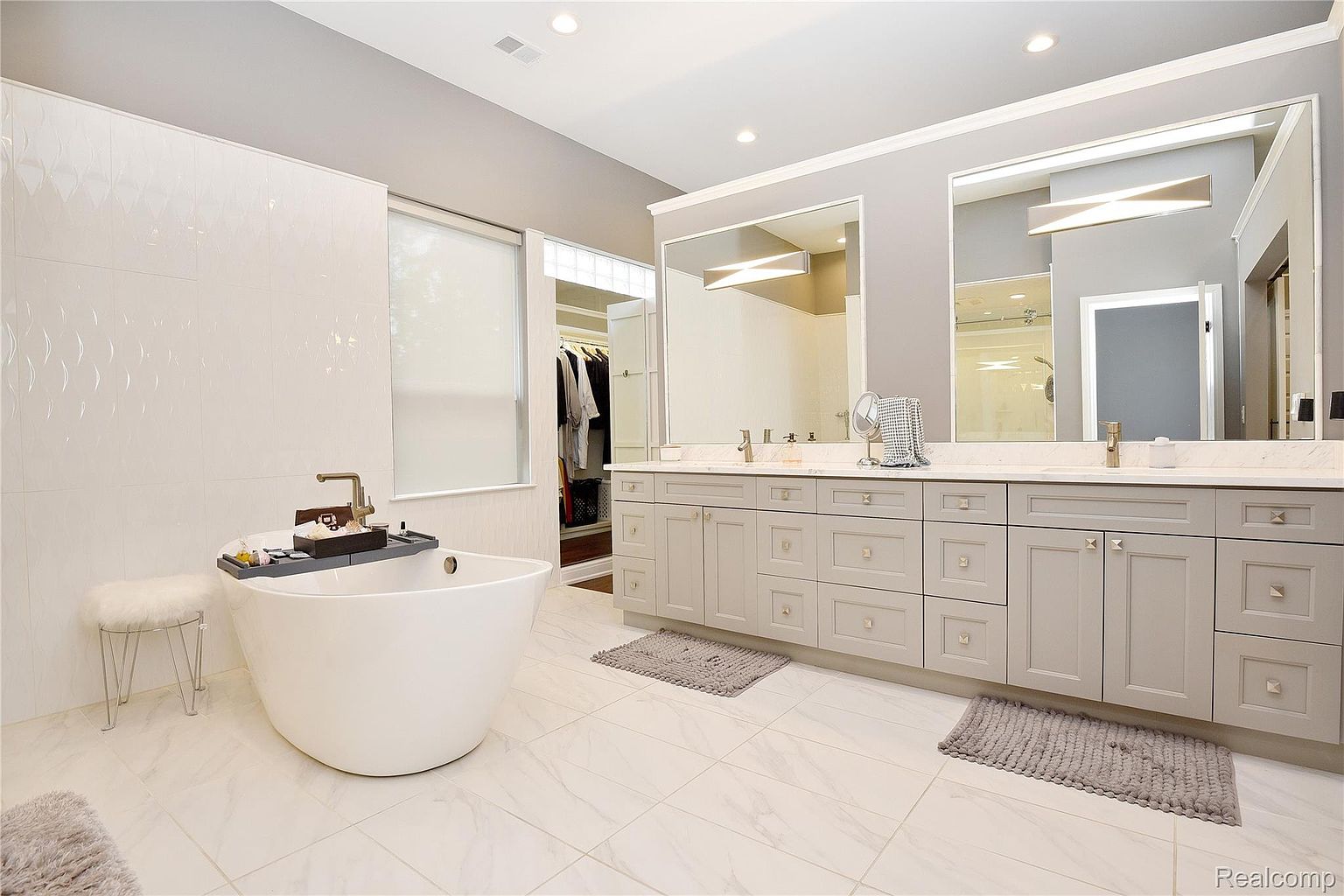 This is a luxurious primary bathroom featuring a freestanding soaking tub with a modern faucet and a tray holding bath accessories. A double vanity with light gray cabinetry, marble countertops, and large mirrors is visible, along with neutral-toned tile flooring and a glimpse into a walk-in closet. The overall impression is clean, bright, and upscale.