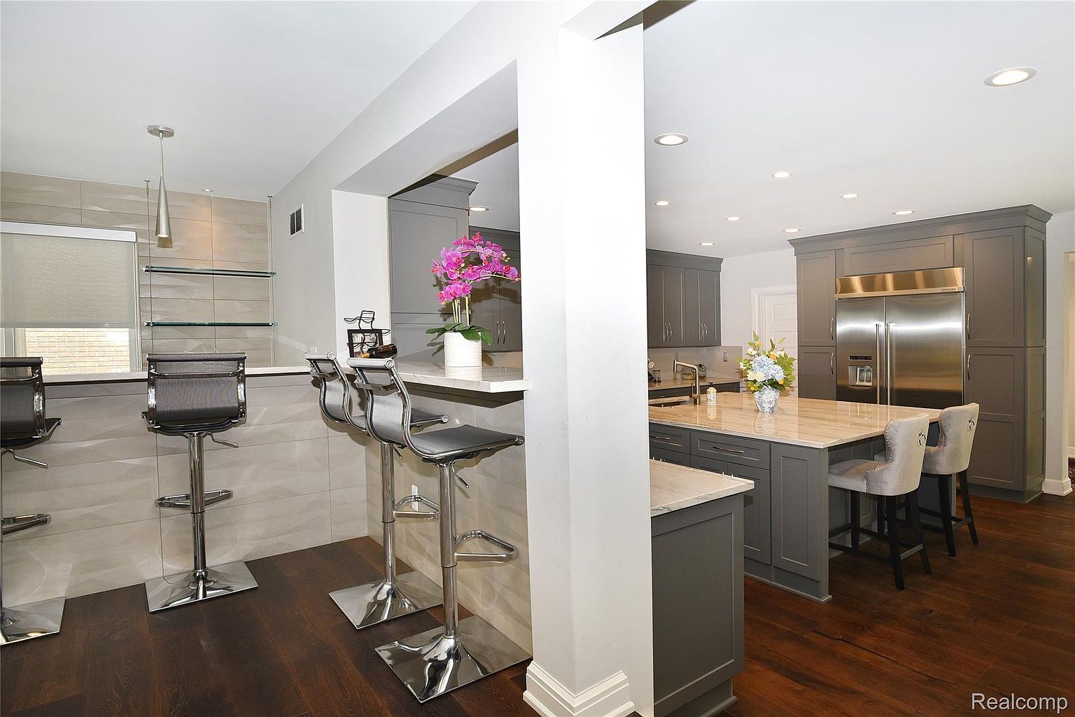 This is a well-lit kitchen featuring gray cabinetry, stainless steel appliances, and a contrasting countertop. A kitchen island with bar seating is present, along with additional bar seating at a counter. The dark wood flooring adds warmth to the space.